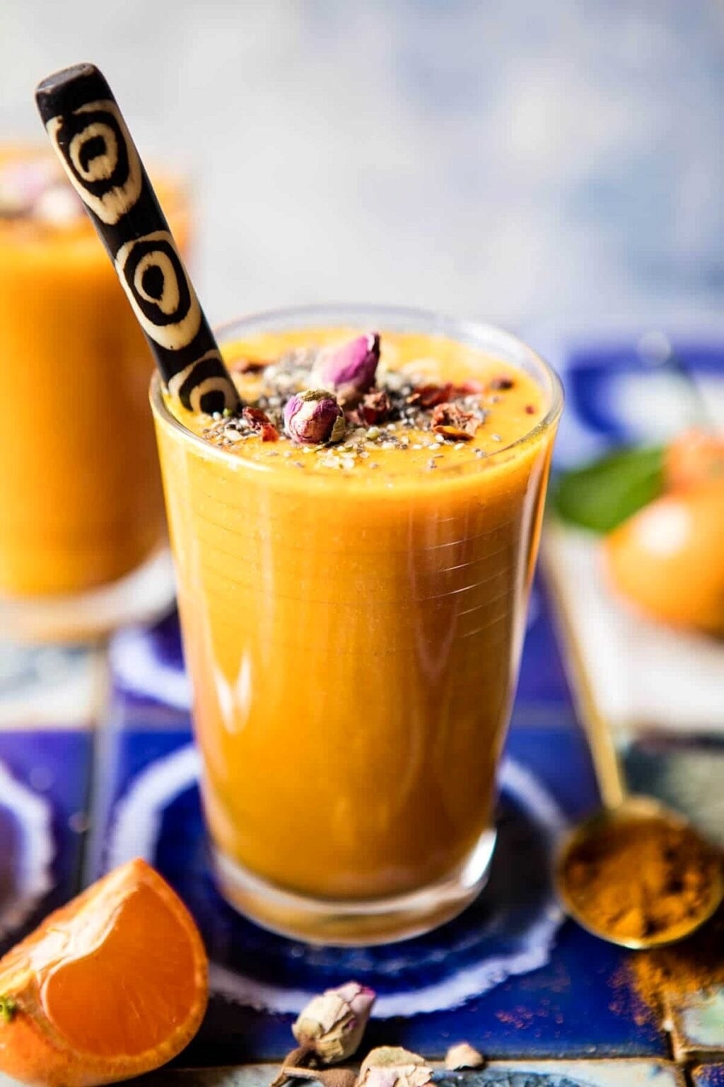 A glass of smoothie garnished with nuts and seeds, with a patterned straw, accompanied by fresh ingredients and a spoon of spices