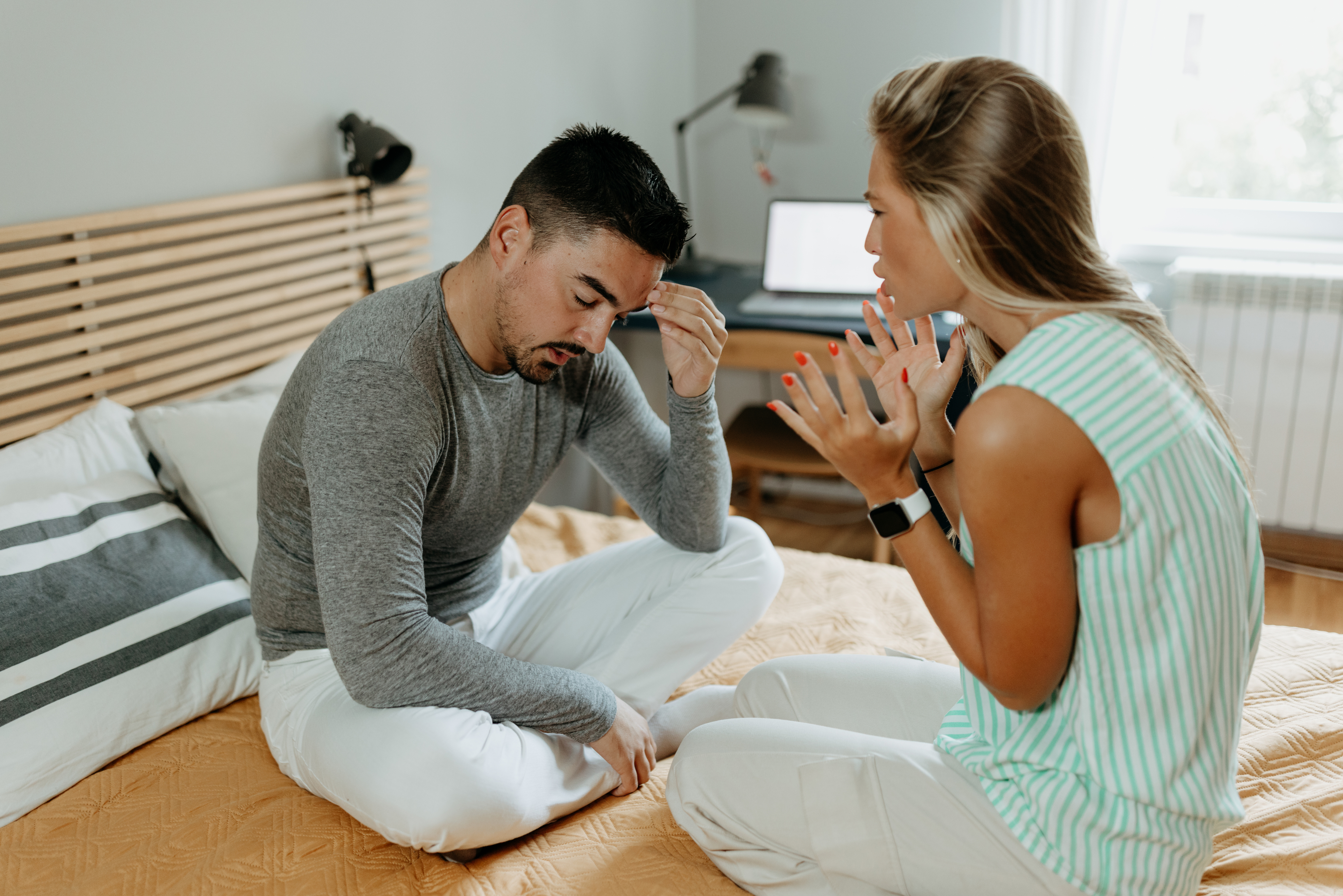 Two people sitting on a bed in a discussion, with one person gesturing and the other looking distressed