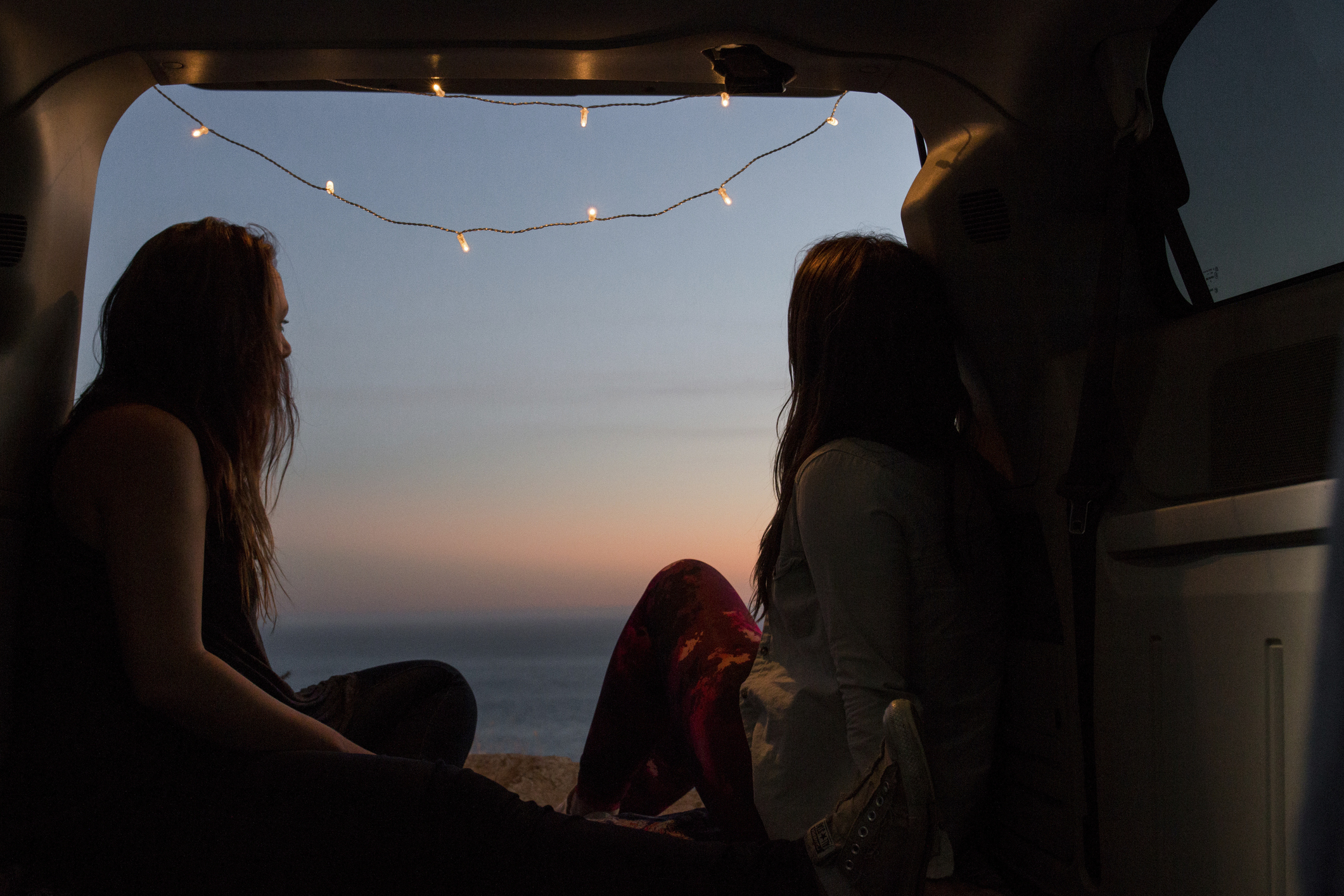 Two people sitting in a van&#x27;s open trunk, facing a sunset, with a string of lights above