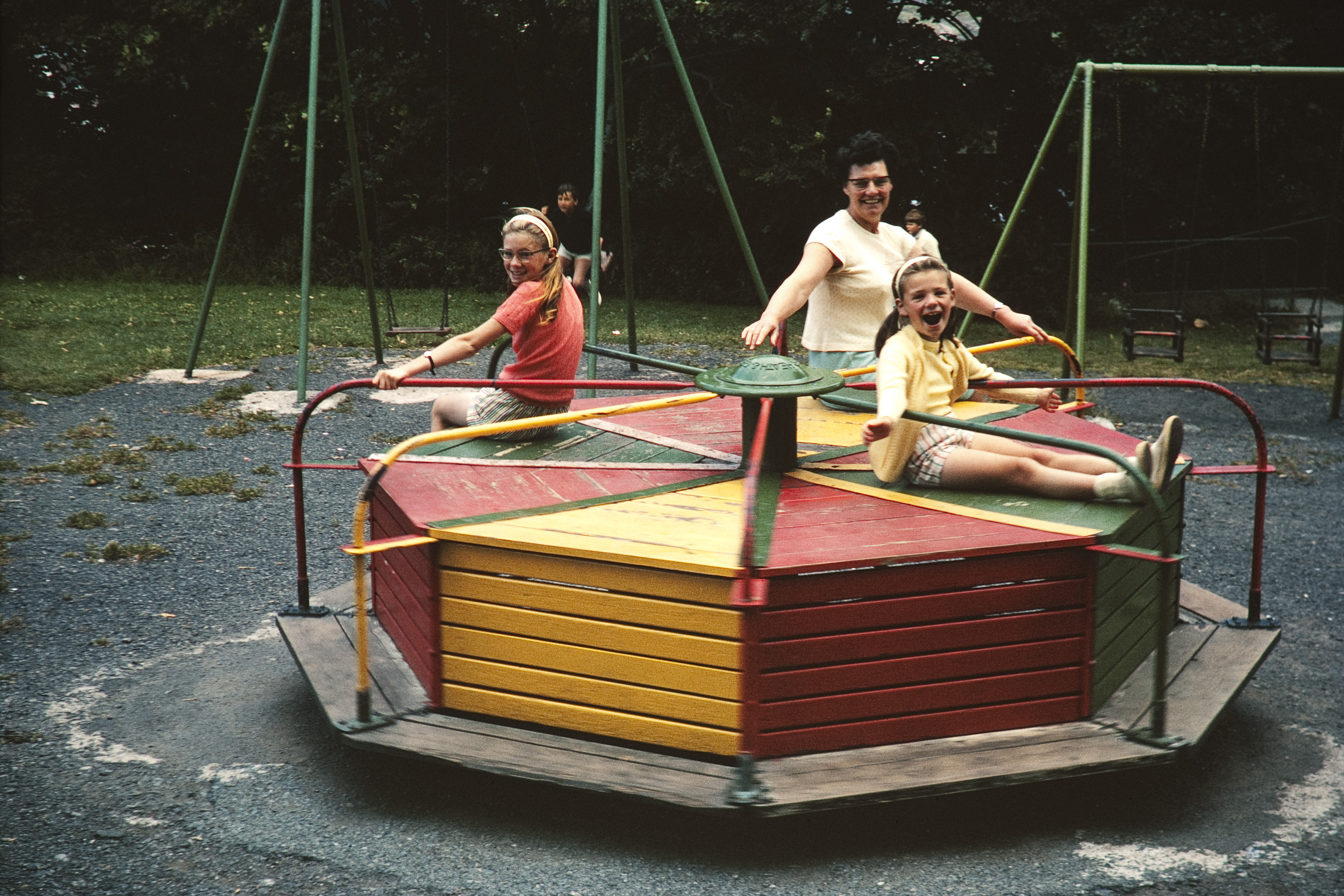 Adult and two children enjoy a merry-go-round at a playground, expressing joy