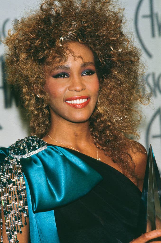Whitney Houston smiling, wearing a teal dress with a bejeweled shoulder, holding an award