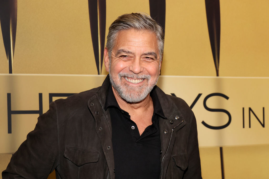 George Clooney smiling in a casual black shirt at an event with a golden background