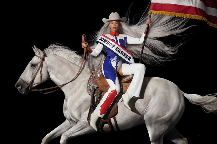 Performer in a cowboy-themed outfit riding a horse, carrying an American flag, representing a music event or performer