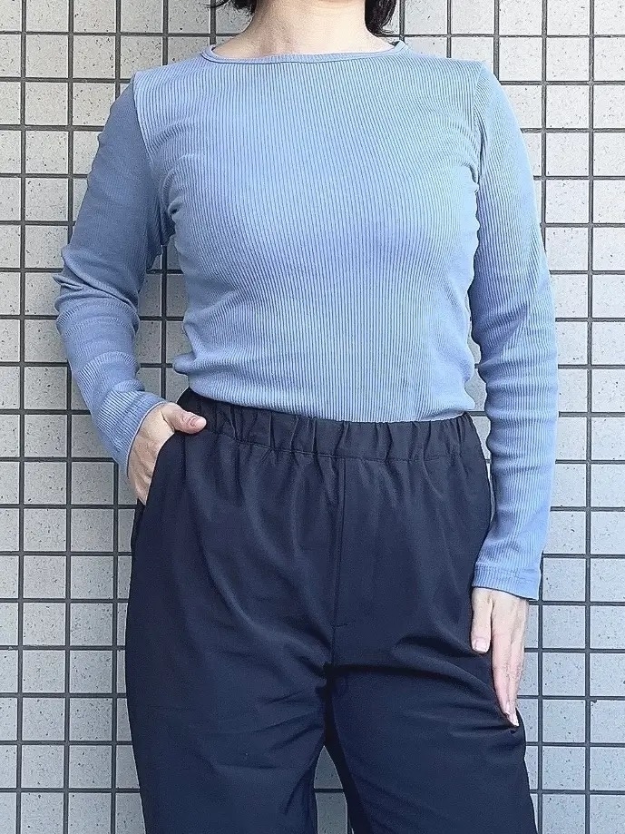 Person in a blue ribbed top and black pants, standing against a tile wall, face not visible