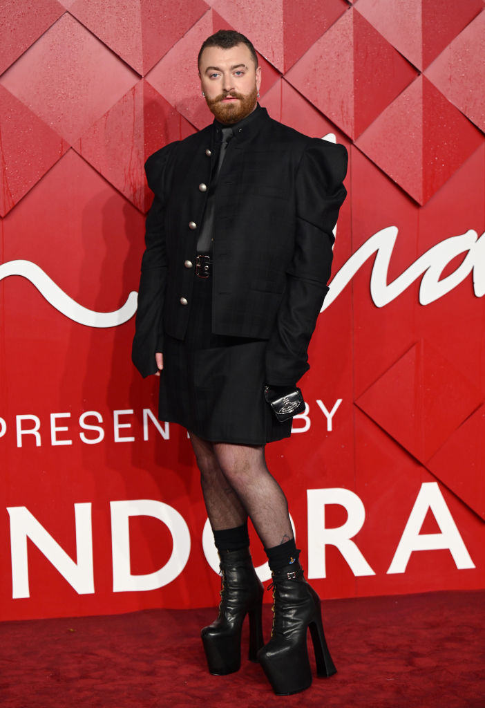 Sam Smith standing on the red carpet wearing a black jacket with oversized sleeves, a matching skirt, and platform boots
