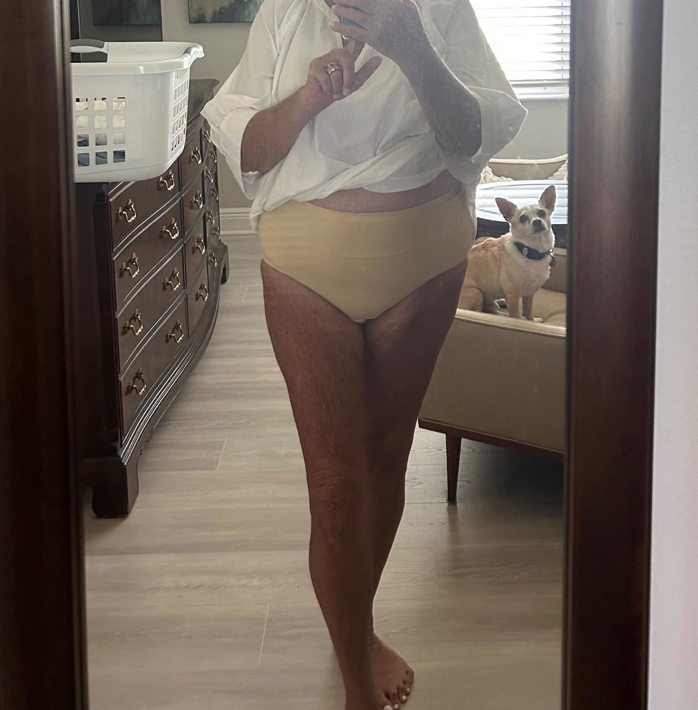 Person in beige underwear and white top standing in front of a mirror with a dog on the bed in the background