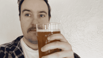 Man holding a glass of beer, wearing a plaid shirt, and smiling at the camera