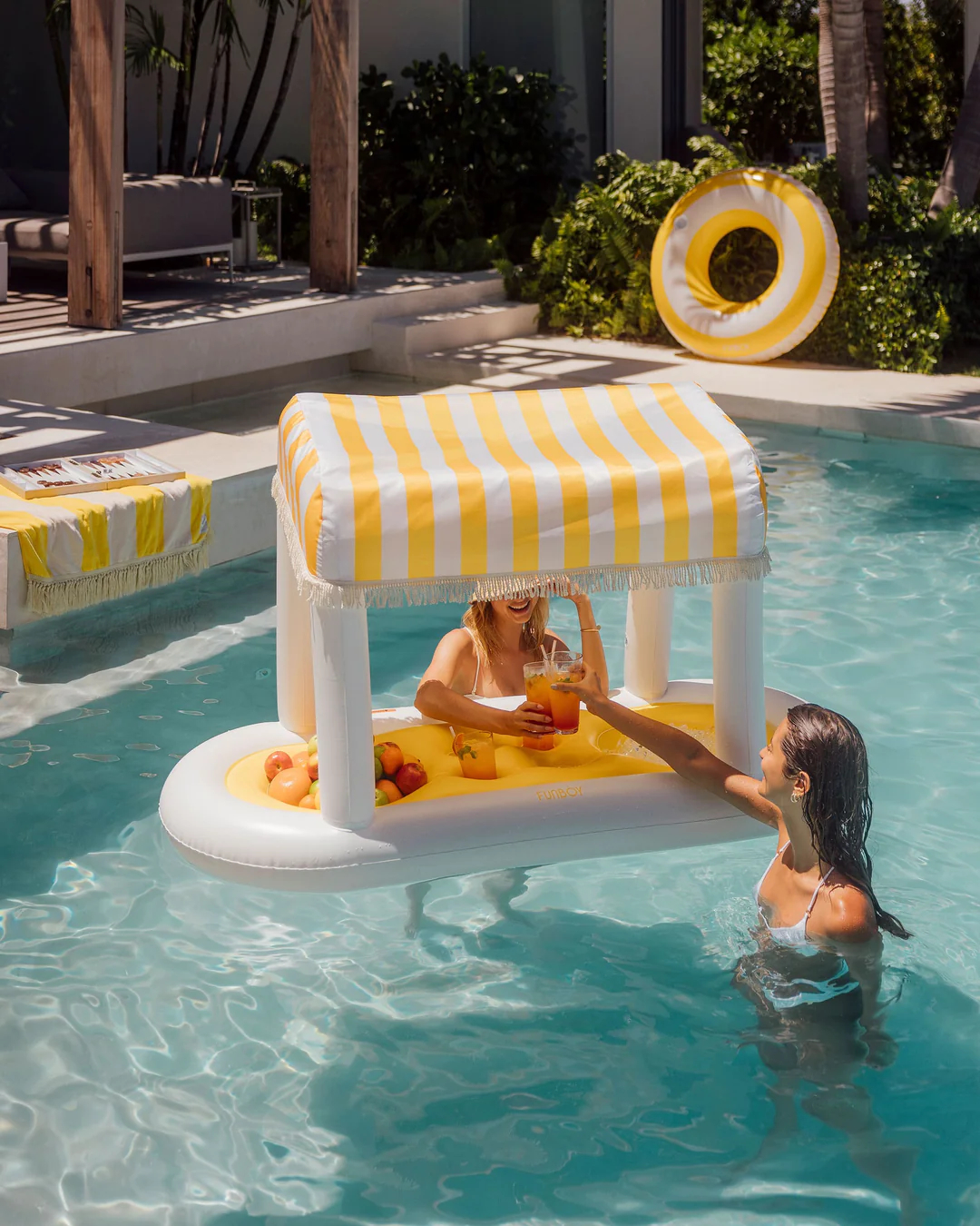 Two people cheersing in a pool with a striped canopy float filled with fruit