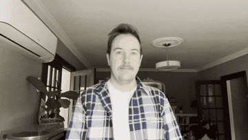 Man in a plaid shirt standing in a room with a ceiling fan and plant in the background