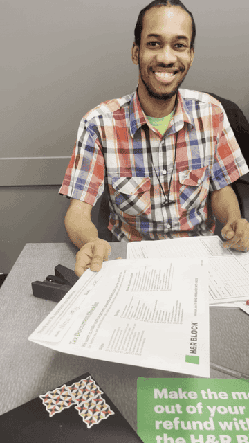 Un homme vêtu d'une chemise à carreaux, assis à un bureau avec des documents et souriant devant la caméra.