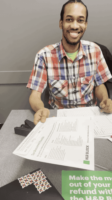 Un homme vêtu d&#x27;une chemise à carreaux, assis à un bureau avec des documents et souriant devant la caméra.