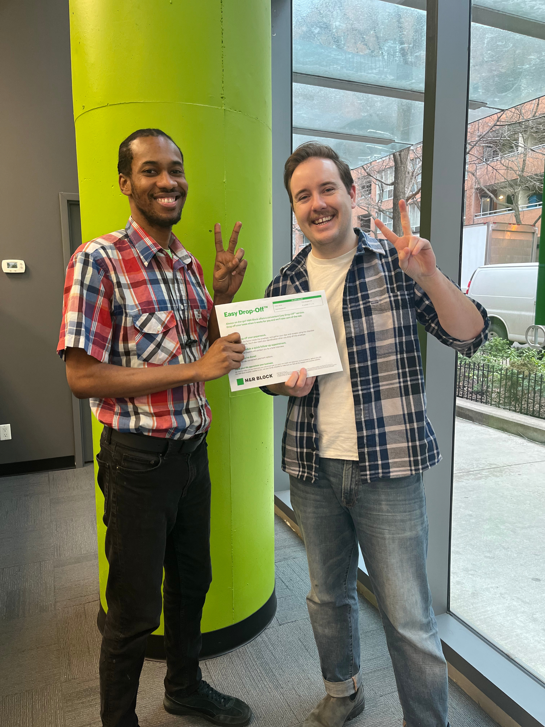 Two people smiling, one holding a certificate, standing indoors next to a pillar, both making peace signs