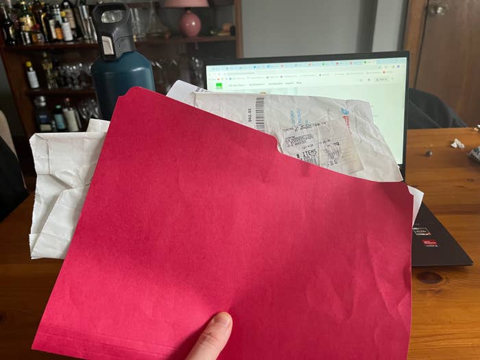 Person holding a red folder over a desk with receipts sticking out, laptop in the background