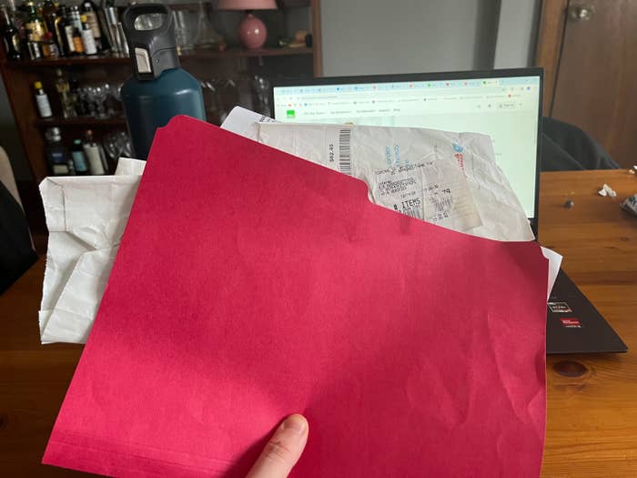 Une personne qui tient un classeur rouge au-dessus d'un bureau avec des reçus qui y sont collés, un ordinateur portable à l'arrière-plan.