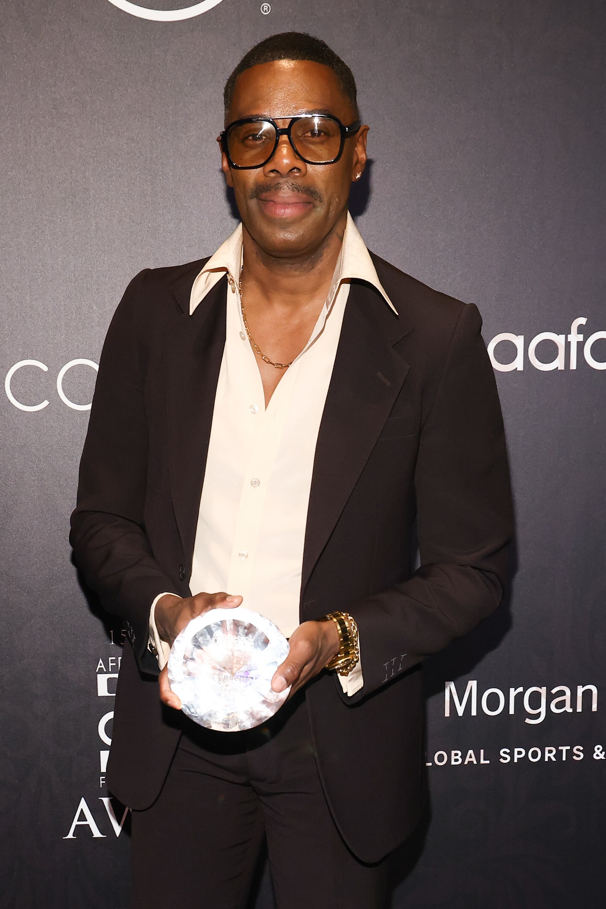 Colman Domingo posing with an award, wearing glasses, and a suit with an unbuttoned shirt