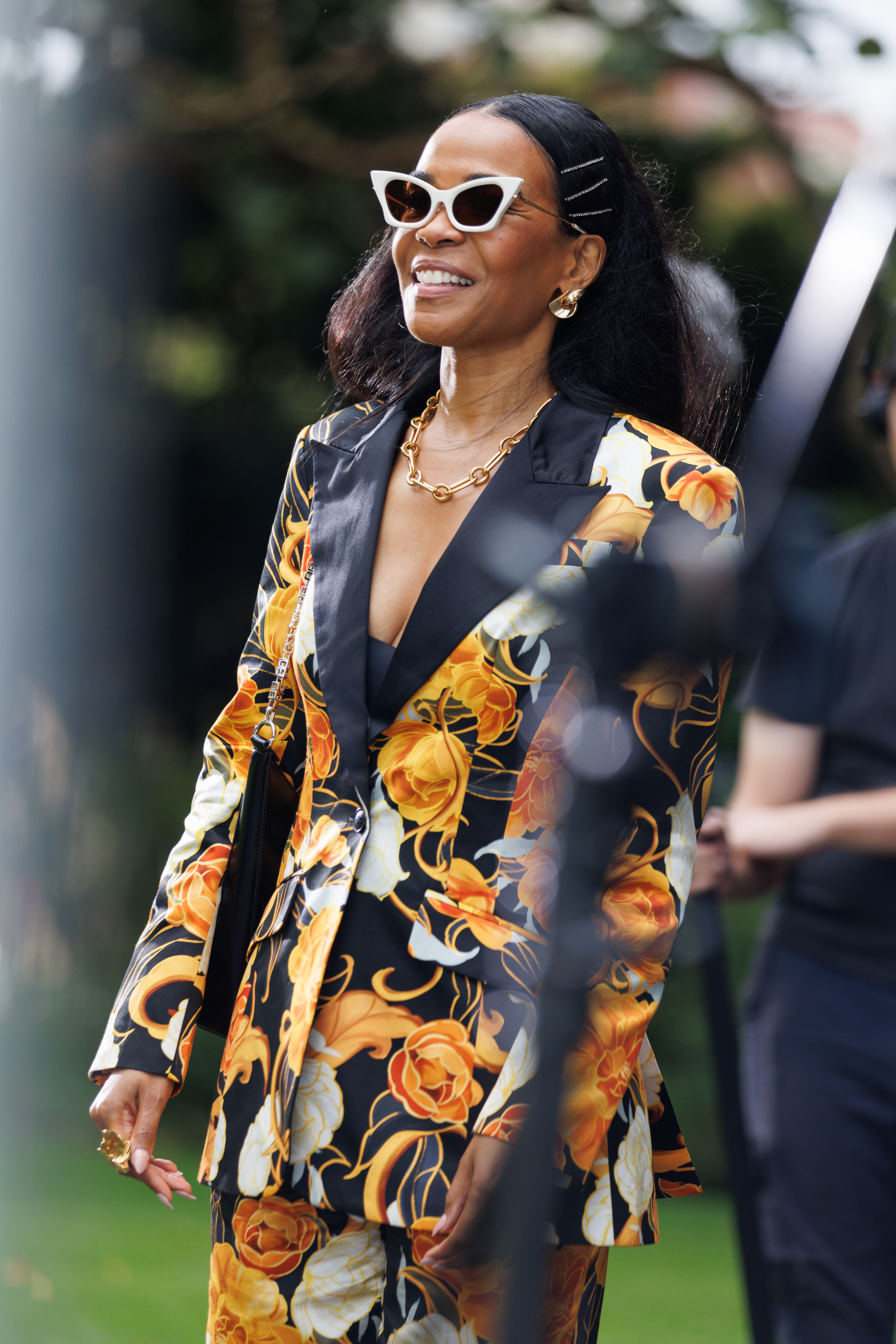 Woman in floral suit with sunglasses and necklace, smiling behind a blurred foreground