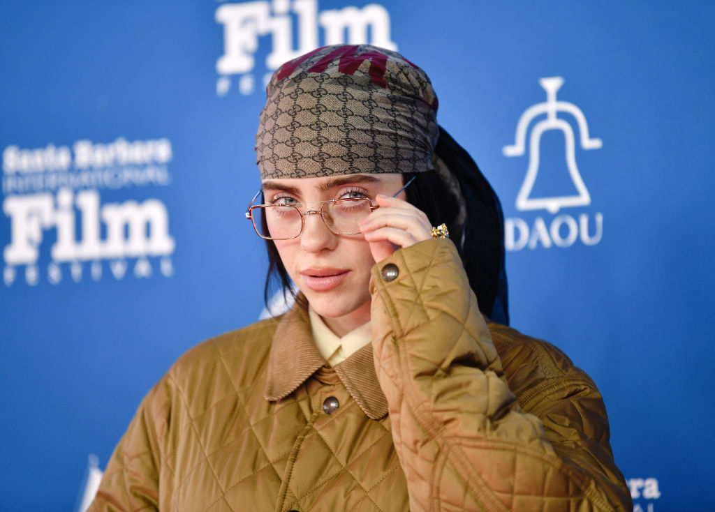 Billie Eilish at an event wearing a patterned headscarf, large glasses, and a quilted jacket