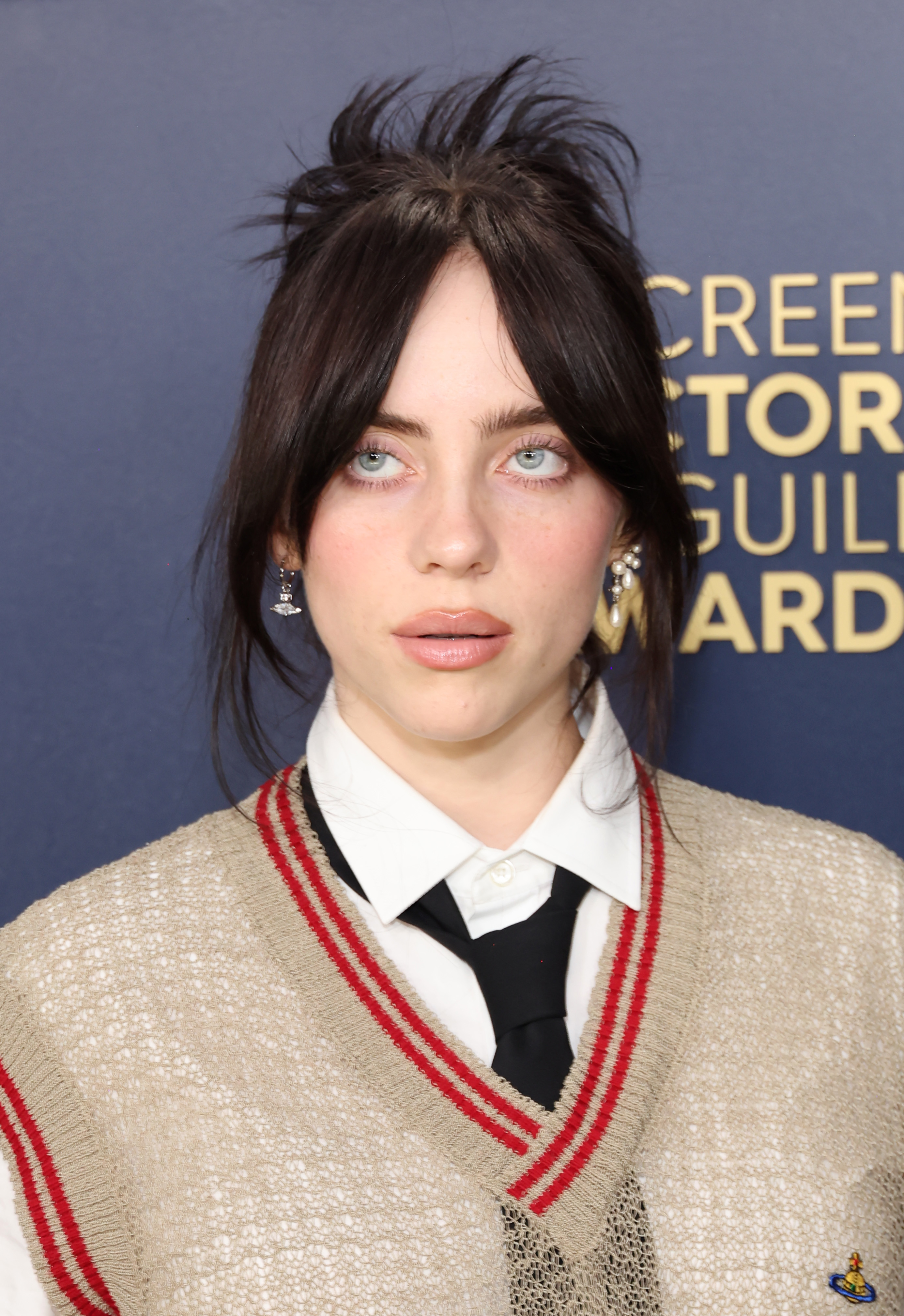 Billie Eilish at an event wearing a beige sweater vest over a white shirt with lace detailing