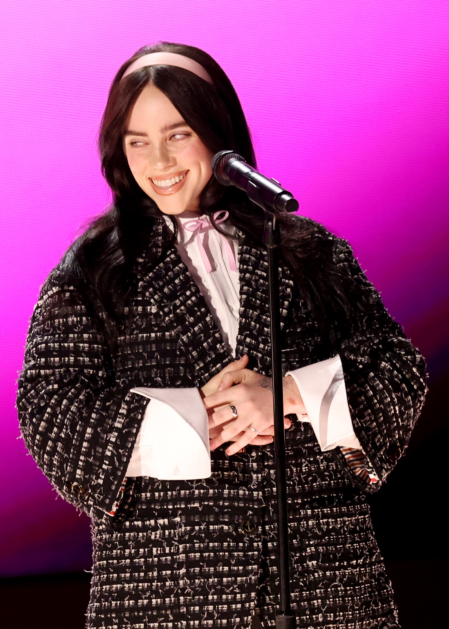 Billie Eilish stands at a microphone in a textured coat and ribbon-tied blouse, smiling
