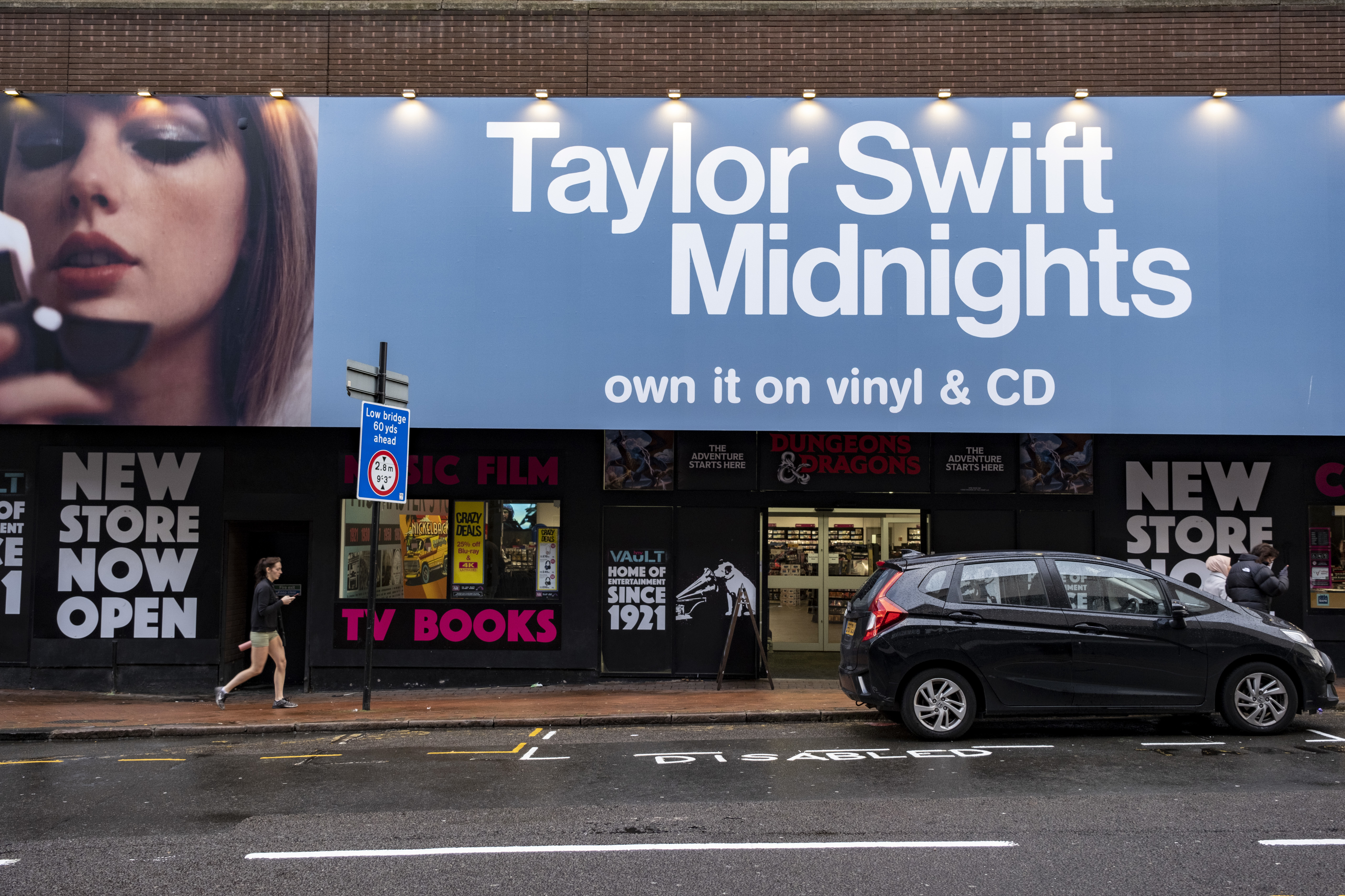 Billboard featuring Taylor Swift advertising her &quot;Midnights&quot; album available on vinyl and CD above a street scene with shops and a car