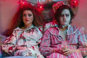 Lisa and the Creature from "Lisa Frankenstein" in patterned pajamas sitting beside each other looking distressed, in a dimly lit room with string lights