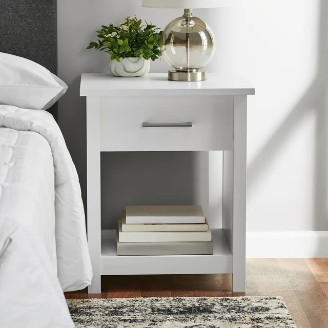 White nightstand with a drawer, a gold lamp on top, and books underneath, beside a bed