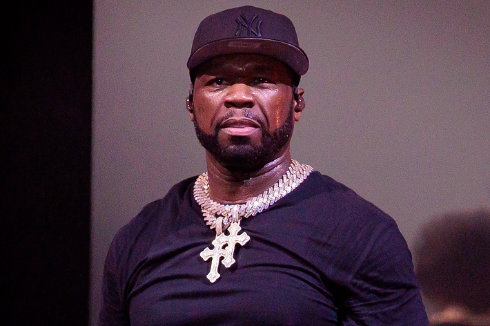 Person on stage wearing a cap, black top, and a large cross necklace; appears focused