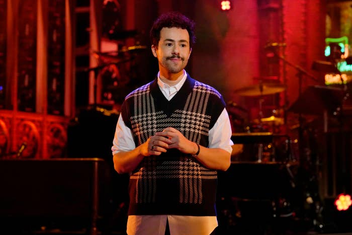 A person on stage wearing a patterned vest and white shirt with hands clasped standing in front of a microphone