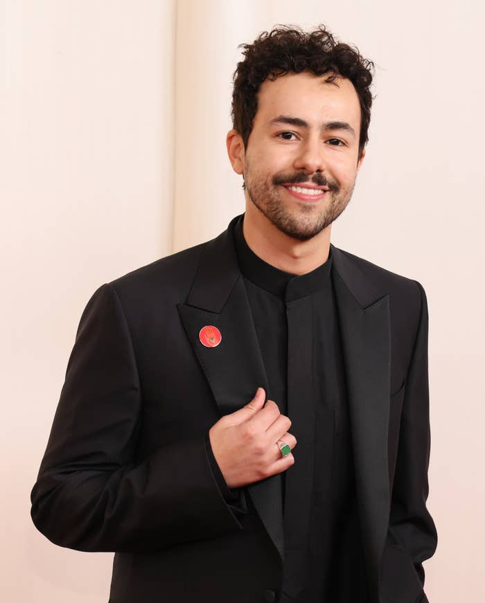 Man in black suit with lapel pin and ring, posing with hand on chest, smiling
