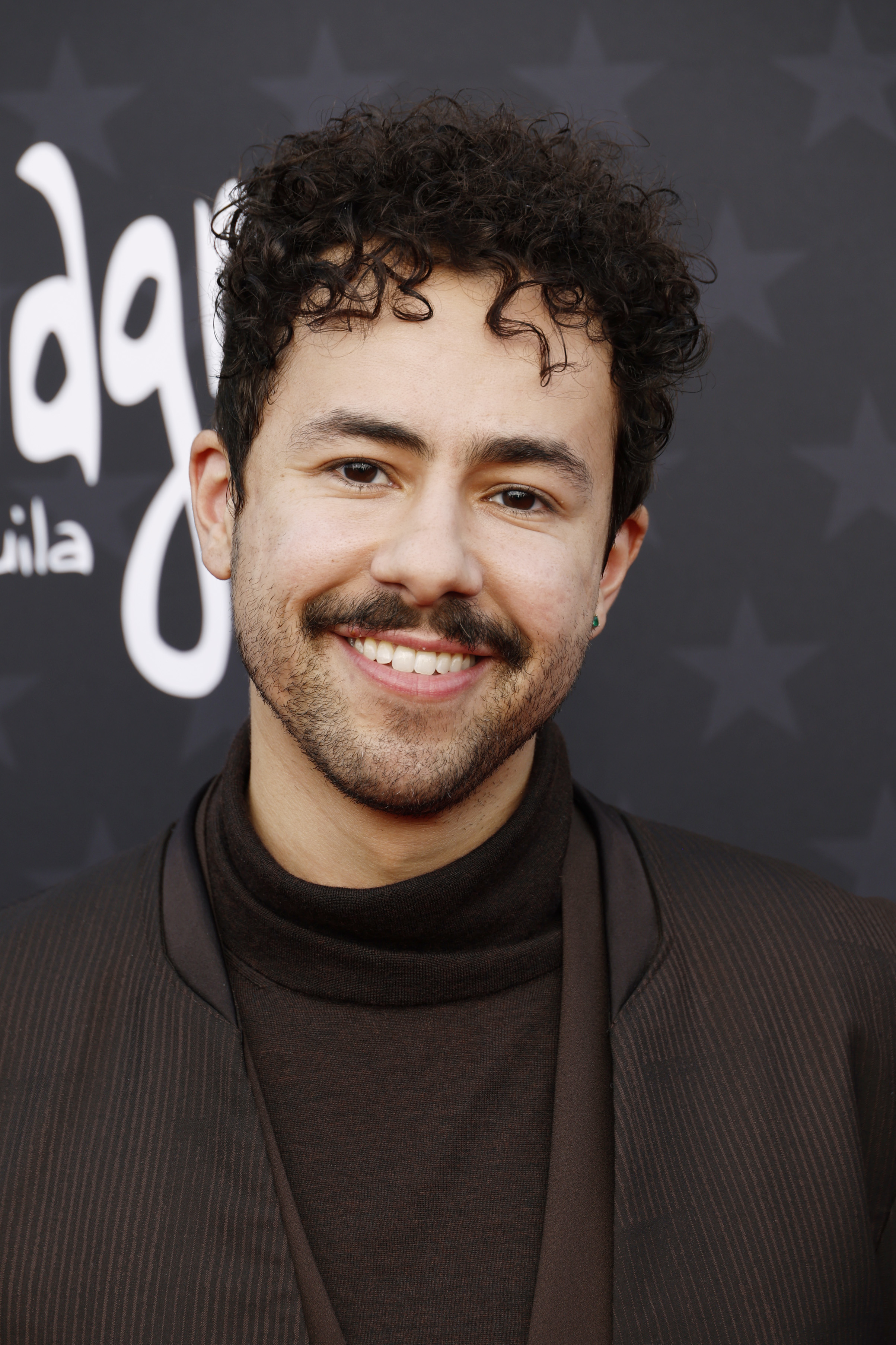 Man with curly hair smiling, wearing a high-neck top and jacket at an event