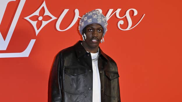 Lil Yachty in a leather jacket and sparkling head accessory stands before a logo backdrop