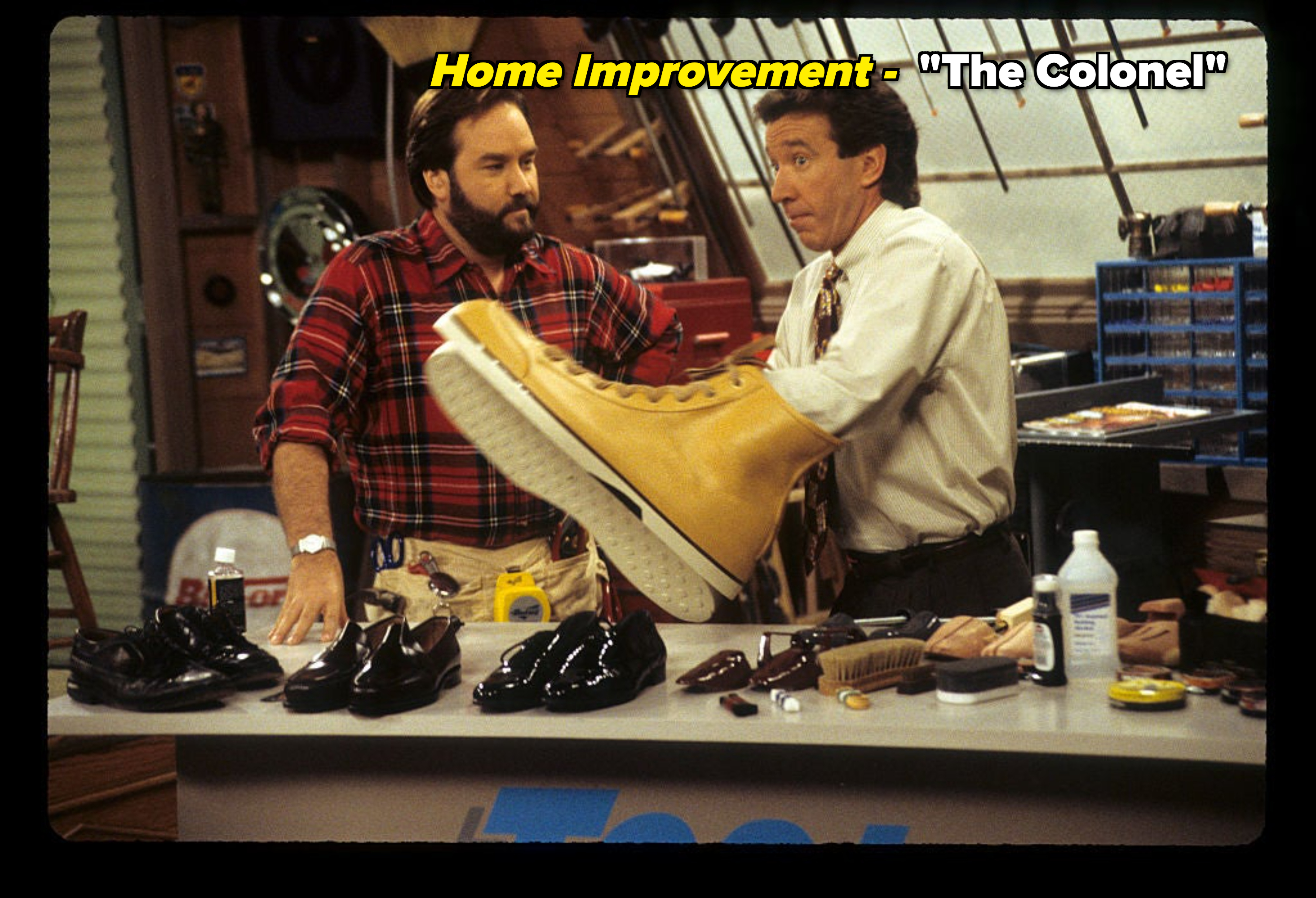 Two men on a TV show set, one holding a large boot, surrounded by various shoes and tools