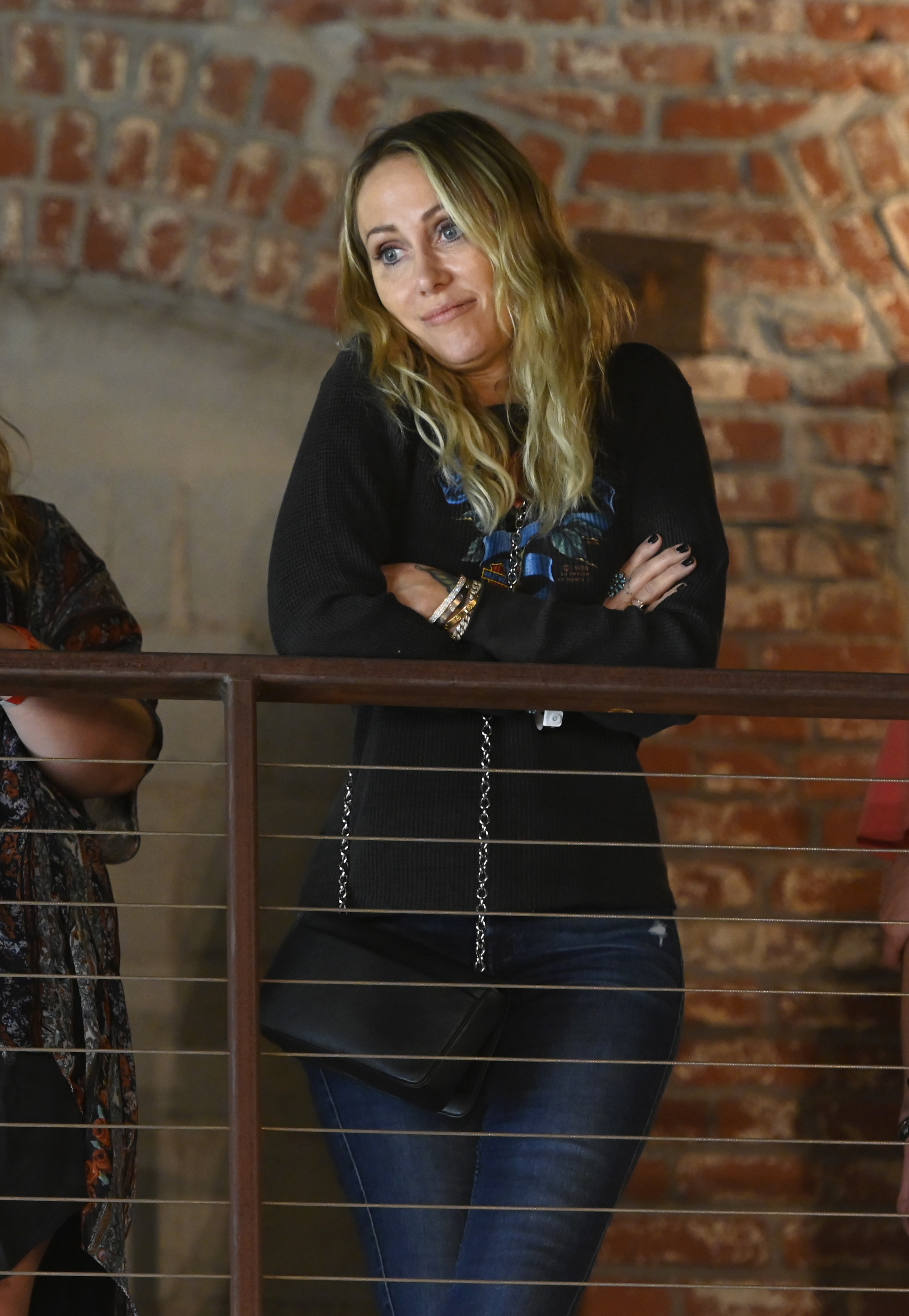 Woman in a black sweater and jeans leaning on a railing indoors, looking to the side with a slight smile