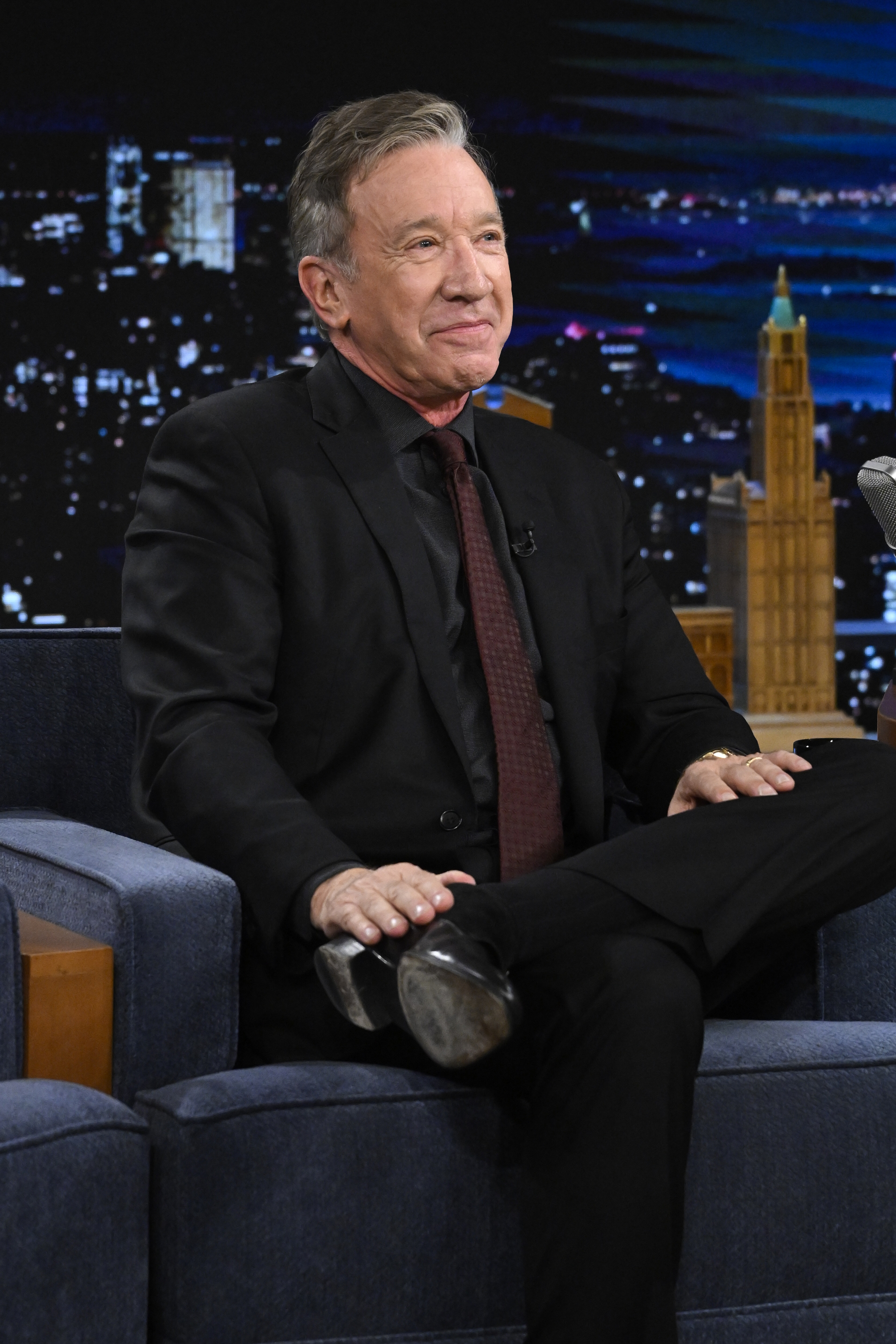Person seated on a talk show set, wearing a black suit and looking off-camera with a relaxed expression