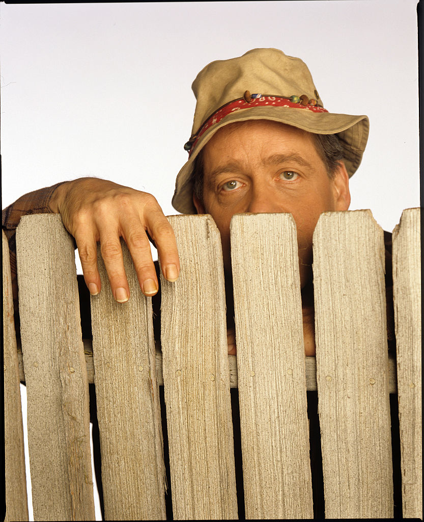 Man in bucket hat peeking over a wooden fence, only eyes and top of face visible