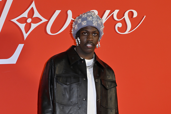 Lil Yachty in a leather jacket and sparkling head accessory stands before a logo backdrop