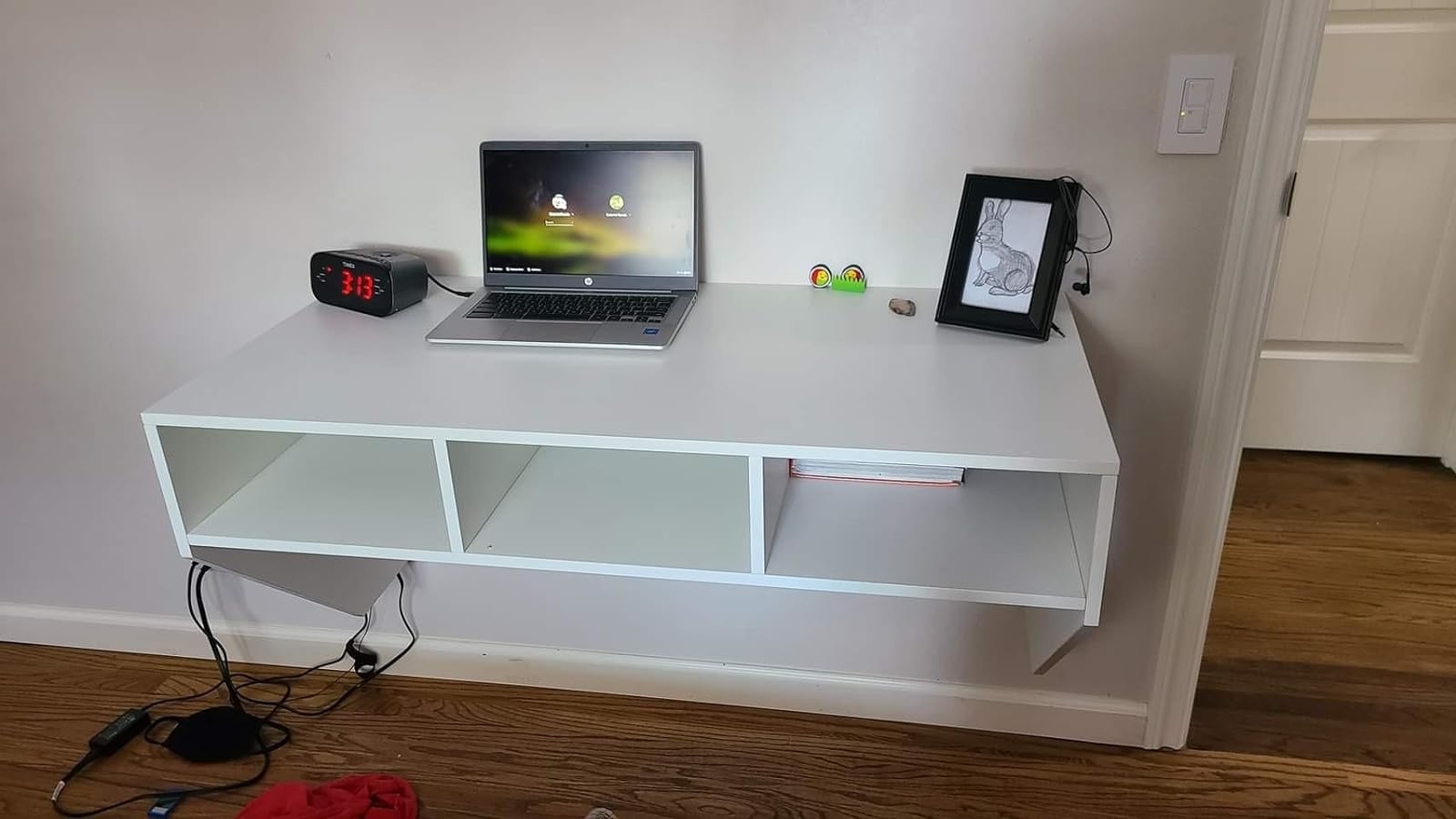 White minimalist shelf with laptop, digital clock, and framed photo in a domestic setting, indicating a tidy home office space for online shopping