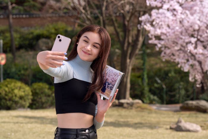 Person taking a selfie with a book, standing near blooming trees