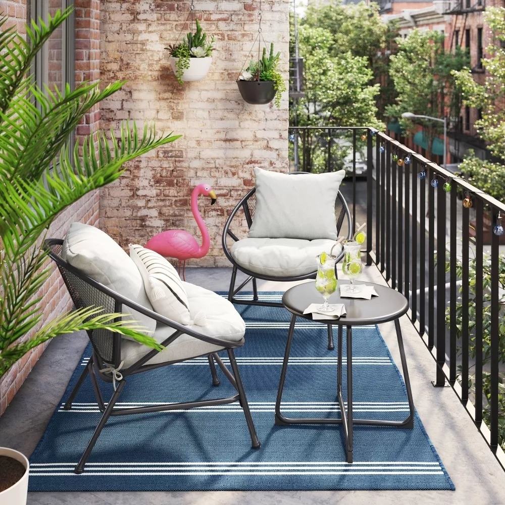 A cozy balcony setup with two chairs, cushions, a side table with drinks, plants, and a decorative flamingo