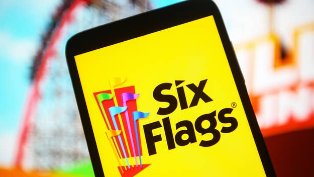 Smartphone with Six Flags logo on screen, partial view of roller coaster and sky in background