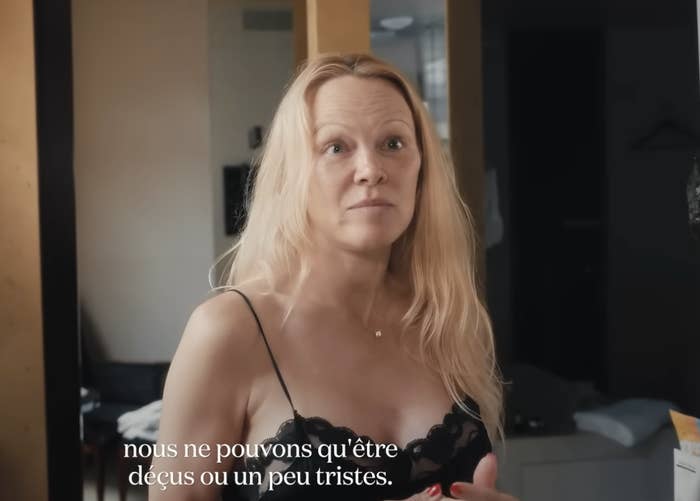 Pamela in a black lace top sits indoors, with French captioning at the bottom