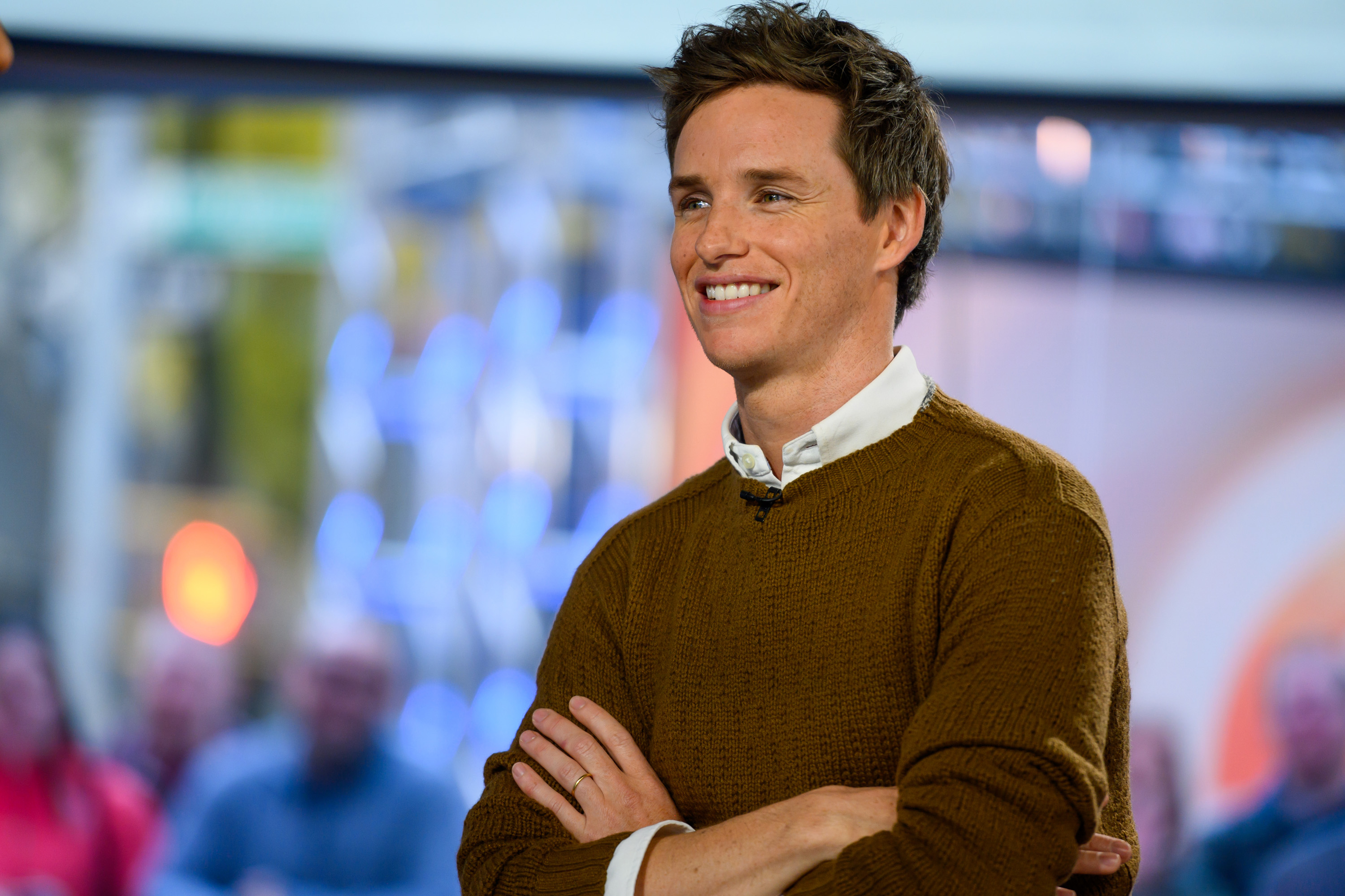 Eddie Redmayne smiling in a studio, wearing a sweater over a collared shirt, arms crossed