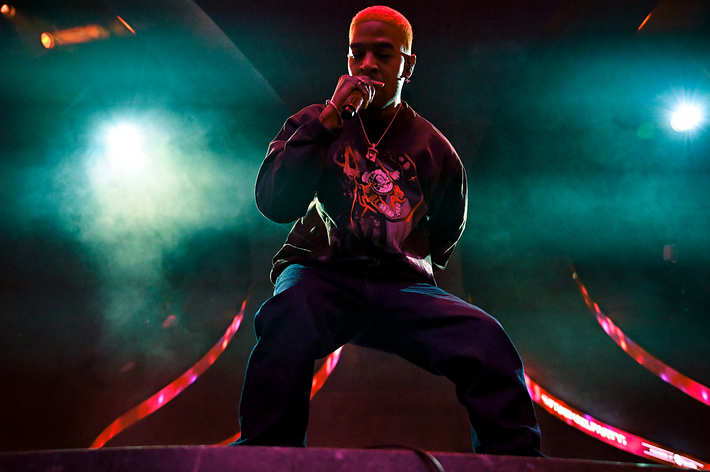 Kid Cudi on stage singing with a microphone, wearing casual attire with graphic design, under spotlights