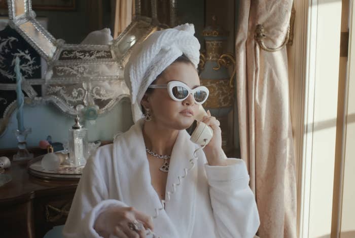 Selena Gomez in a music video scene, wearing a bathrobe and sunglasses, sitting by a window with a view and talking on a landline phone