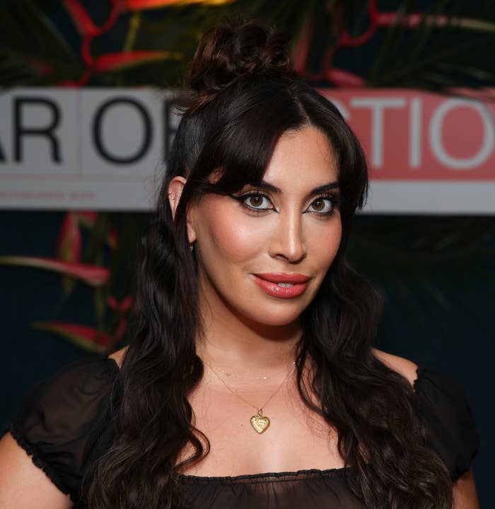 A closeup of Sarah Bahbah whose hair is styled in a half-updo and is pendant necklace standing before a 'Year of Emotion' backdrop