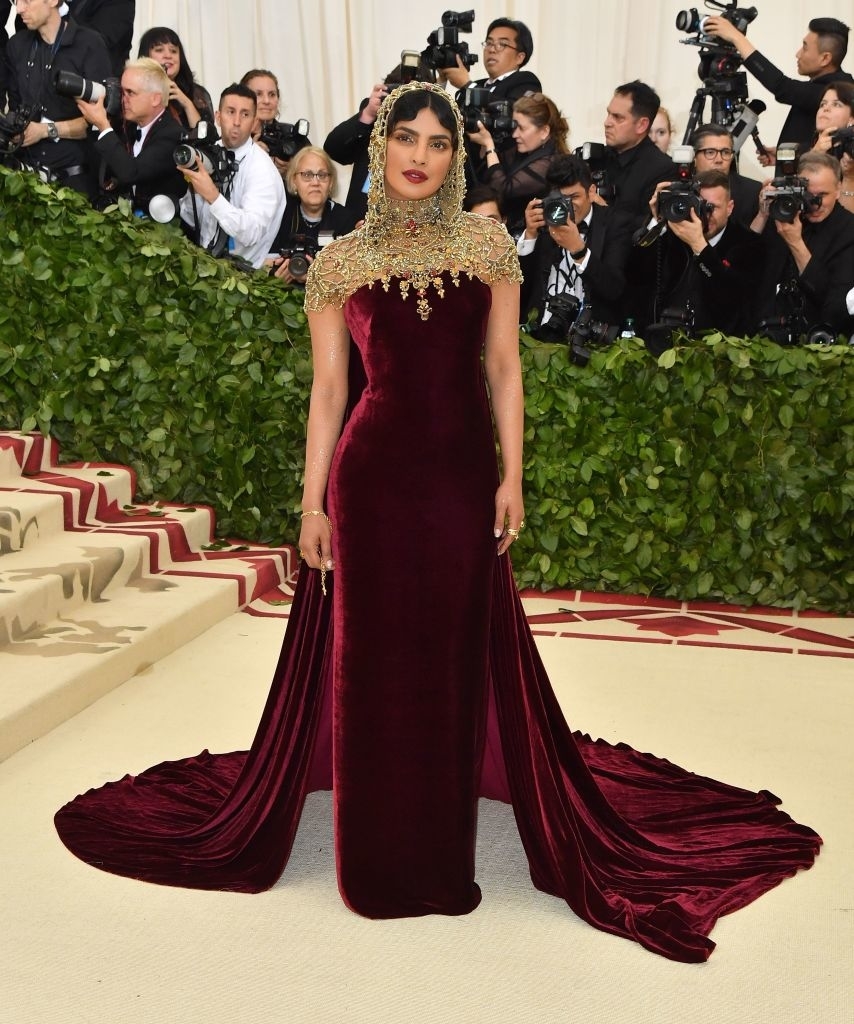 Priyanka in a velvet gown with a gold headpiece, standing on stairs with photographers in the background