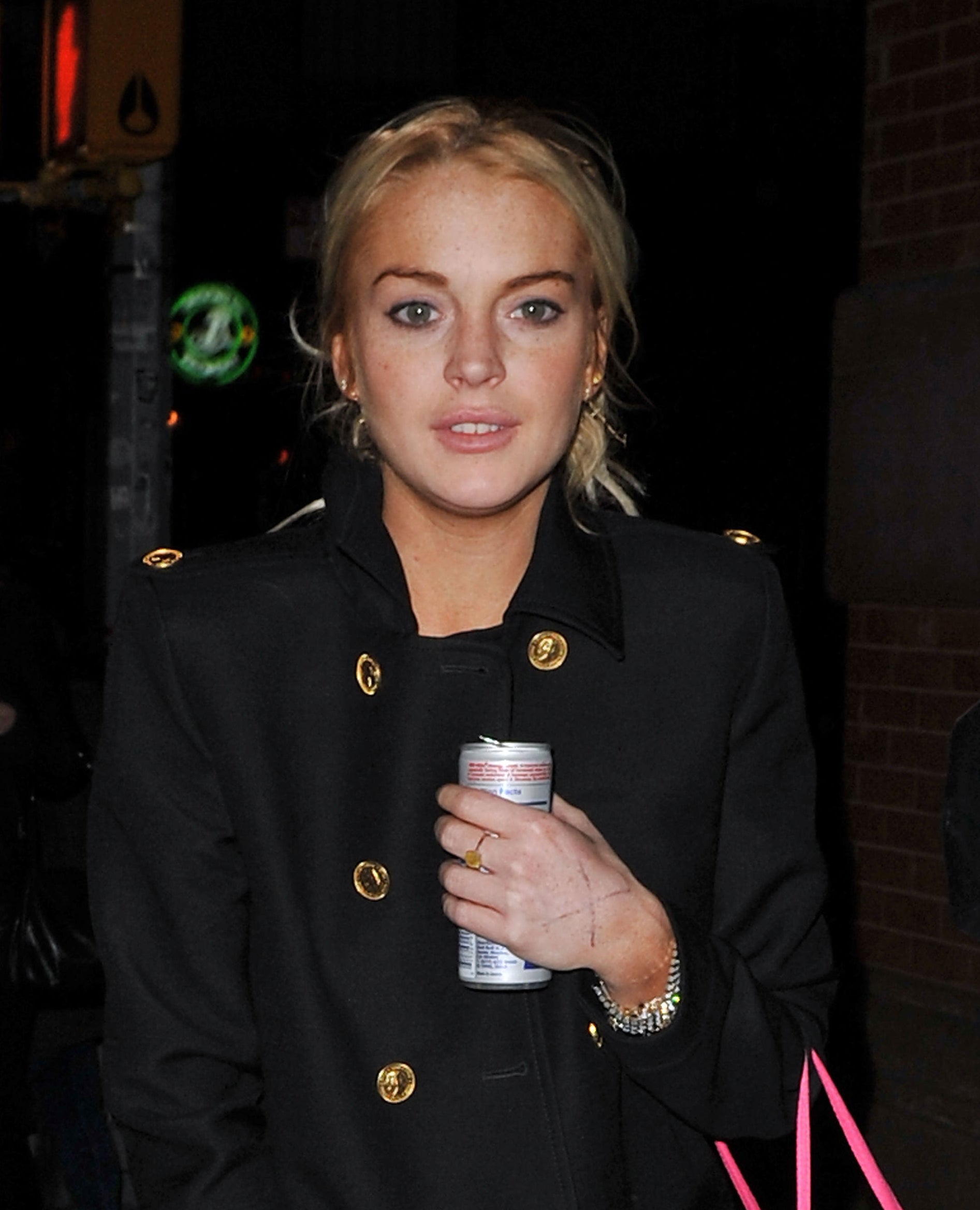Lindsay Lohan in a double-breasted jacket holding a canned drink, standing on a sidewalk at night