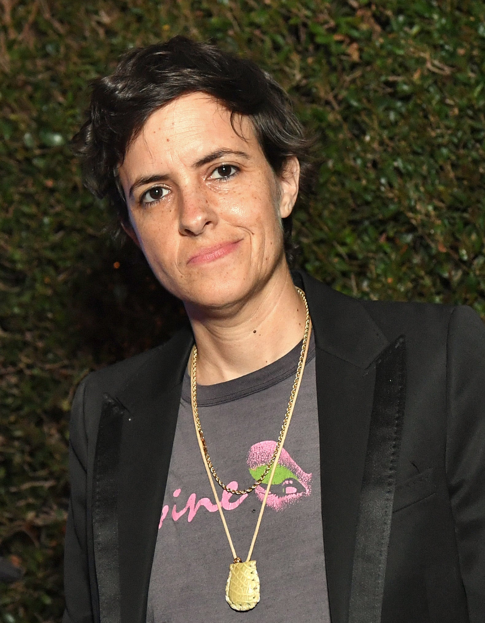 Samantha Ronson with a headset around her neck stands by DJ equipment; wears a black blazer over a graphic tee