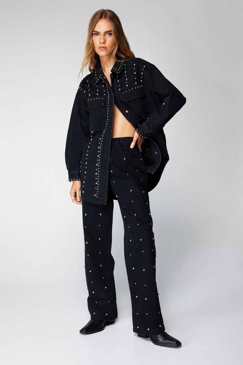 Model in a studded black denim jacket and matching trousers, midriff exposed, hands in pockets