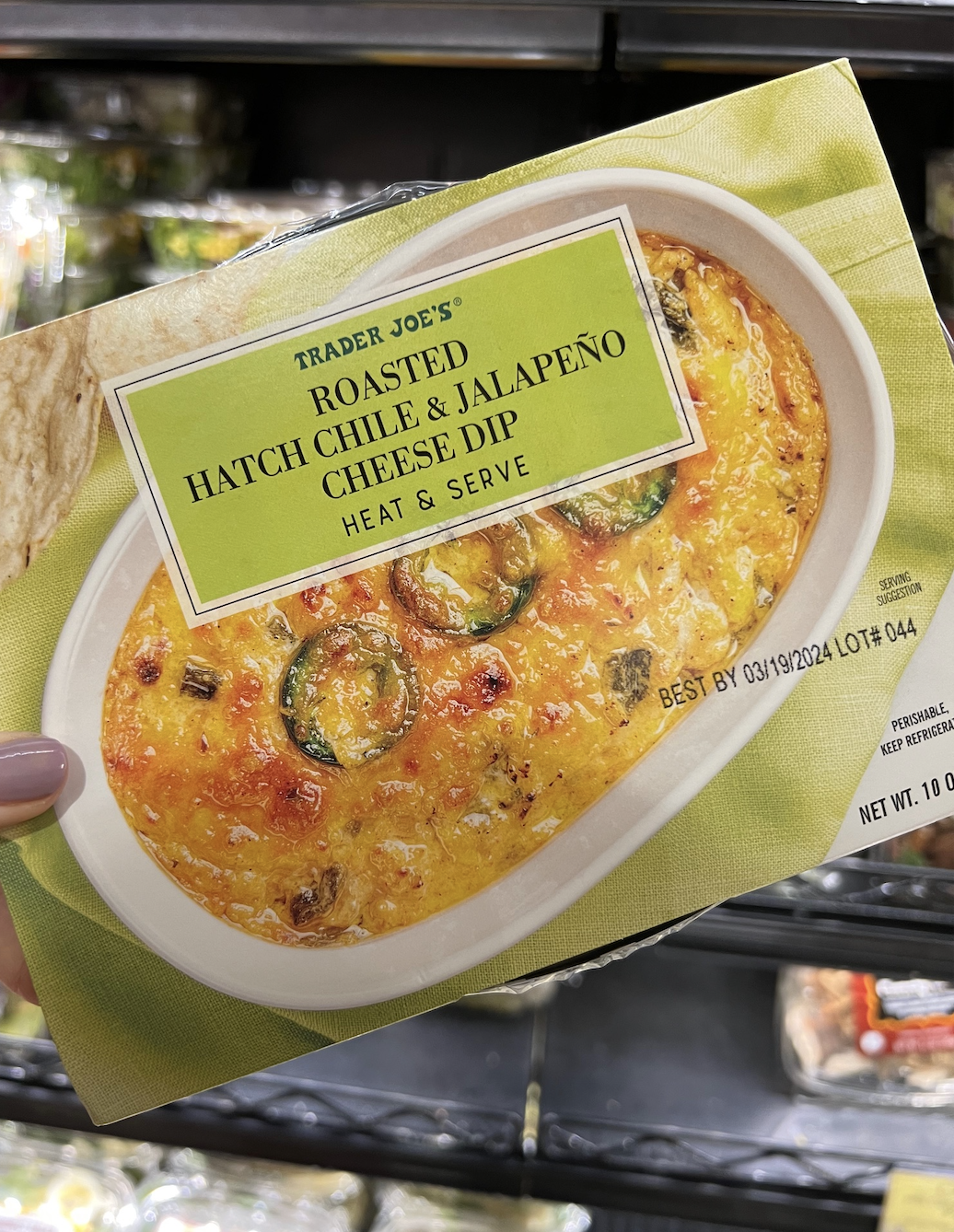 Hand holding a package of Trader Joe&#x27;s Roasted Hatch Chile &amp;amp; Jalapeño Cheese Dip in a grocery store